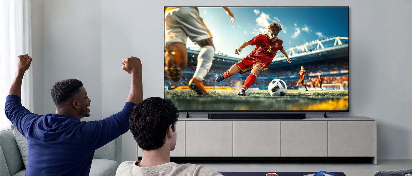 Dw&oacute;ch m&eogon;&zdot;czyzn siedzi w jasnym pokoju i ogl&aogon;da na telewizorze mecz pi&lstrok;karski, gdy jeden z nich podnosi si&eogon; z rado&sacute;ci. 